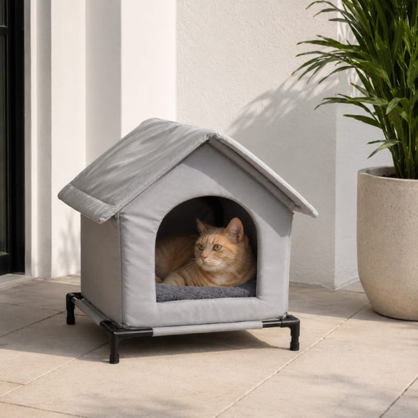 Abri pour chat extérieur gris avec toit souple et base surélevée, placé sur une terrasse lumineuse avec un chat roux à l’intérieur