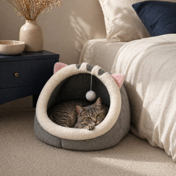 Cocon pour chat gris avec intérieur en peluche douce et pompon suspendu, installé dans une chambre cosy avec un chat allongé dedans
