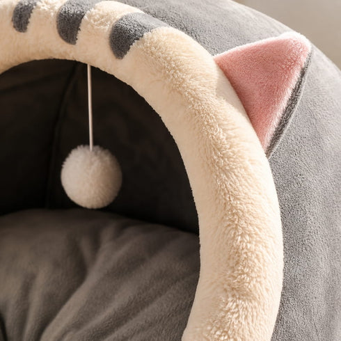 Gros plan du cocon pour chat gris en peluche douce montrant l’entrée rembourrée, l’oreille décorative et le pompon de jeu suspendu.