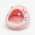Cocon pour chat rose et blanc en peluche avec coussin moelleux, oreilles décoratives, nœud et pompon suspendu sur fond neutre.