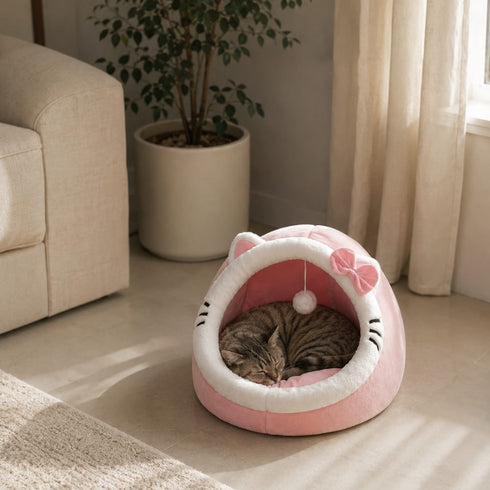 Cocon pour chat rose en peluche douce avec nœud décoratif, installé dans un salon lumineux où un chat dort confortablement à l’intérieur.