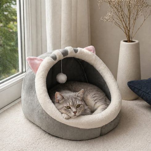 Cocon pour chat en peluche grise et blanche, installé près d’une fenêtre, avec un chat allongé à l’intérieur sur un couchage douillet.