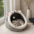 Cocon pour chat en peluche grise et blanche, installé près d’une fenêtre, avec un chat allongé à l’intérieur sur un couchage douillet.