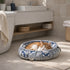 Chat roux et blanc endormi dans un couffin rond gris et bleu à motifs, installé dans une salle de bain lumineuse.