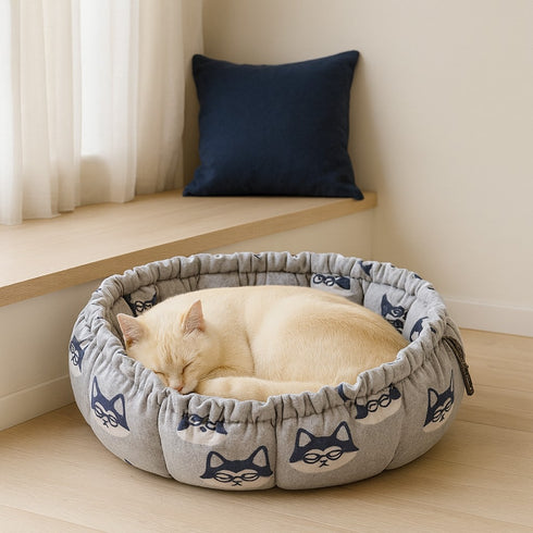 Couffin pour chat en polaire grise avec motifs de têtes de chat bleues, posé près d’une fenêtre, dans lequel un chat crème dort paisiblement.