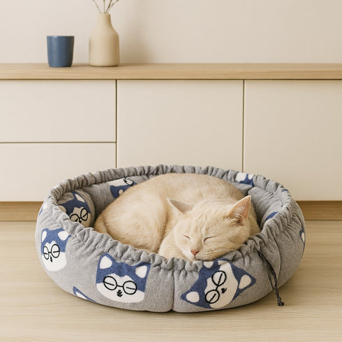 Couffin pour chat en polaire grise avec motifs renard bleus, doté d’un bord froncé, dans lequel un chat beige dort paisiblement posé devant un buffet.