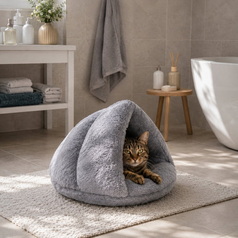 Chat tigré installé dans un coussin chauffant gris en forme de cocon moelleux, posé dans une salle de bain moderne et lumineuse.