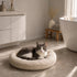 Chat gris et blanc installé dans un coussin pour panier beige en tissu bouclette ultra moelleux, dans une salle de bain moderne et lumineuse.