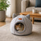 Grotte pour chat grise en tissu doux avec oreilles décoratives, installée dans un salon cosy avec un chat roux à l’intérieur