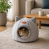 Grotte pour chat grise en tissu doux avec oreilles décoratives, installée dans un salon cosy avec un chat roux à l’intérieur