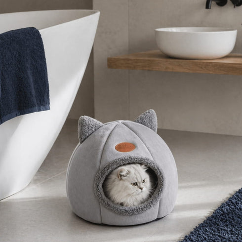 Grotte pour chat grise avec ouverture en peluche et oreilles décoratives, placée dans une salle de bain moderne avec un chat blanc à l’intérieur