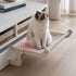 Chat assis sur un hamac de fenêtre sans ventouse rose et gris avec structure en bois installé en intérieur près d’un meuble blanc.