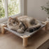 Grand chat allongé sur un hamac pour chat sur pied avec structure en bois naturel et tissu gris quadrillé près d’une fenêtre lumineuse.
