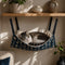 Chat allongé dans un hamac suspendu pour chat en tissu bleu à motifs, accroché sous une étagère pour un espace repos cosy et pratique.