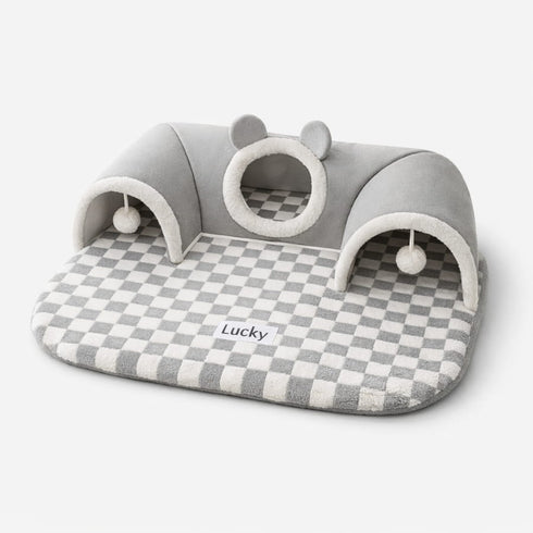 Lit tunnel pour chat gris avec tapis à carreaux, cachettes intégrées et jouets suspendus sur fond studio épuré.