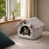 Chat tigré installé dans une maison pour chat grise avec toit en tissu doux, placée près d’une fenêtre lumineuse et apaisante.