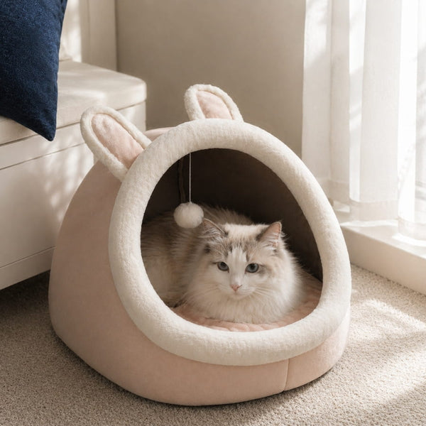 Maison douillette rose pour chat avec oreilles de lapin et intérieur moelleux, installée près d’une fenêtre lumineuse avec un chat allongé à l’intérieur