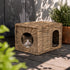 Petite niche pour chat rectangulaire en osier beige avec double ouverture, posée sur une terrasse extérieure avec un chat à l’intérieur