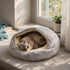 Chat allongé dans un nid moelleux gris clair en fausse fourrure, placé près d’une fenêtre lumineuse dans un intérieur cosy et apaisant.