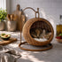Chat allongé dans un panier chat boule osier suspendu en imitation osier avec coussin moelleux, installé dans une cuisine lumineuse et chaleureuse.