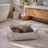 Chat tigré allongé dans un panier pour chat à carreaux gris, installé sur le sol d’une salle de bain lumineuse et chaleureuse