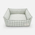 Panier pour chat à carreaux vert avec coussin rayé moelleux et rebords rembourrés sur fond neutre.