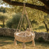 Panier pour chat suspendu en macramé beige avec coussin moelleux, accroché à un arbre pour offrir un espace repos bohème et confortable.