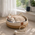 Chat crème allongé dans un panier en rotin pour chat en imitation rotin blanc avec corde de jeu et coussin bleu, placé près d’une fenêtre dans une ambiance douce et lumineuse.