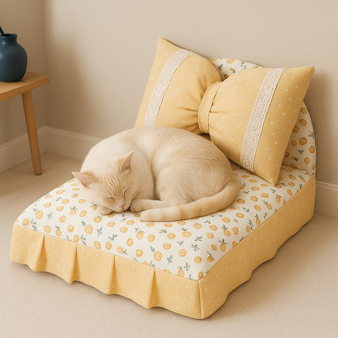 Petit lit pour chat beige à motifs agrumes avec grand coussin nœud, posé au sol dans un intérieur cosy, avec un chat blanc endormi confortablement.