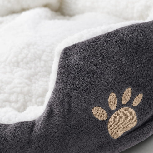 Gros plan sur la texture douce d’un petit panier pour chat gris avec intérieur moelleux blanc et motif patte brodé beige.