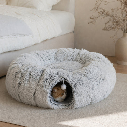 Tanière pour chat grise en peluche douce avec tunnel intégré et pompon suspendu, installée dans une chambre cosy avec un chat roux caché dedans