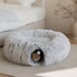 Tanière pour chat grise en peluche douce avec tunnel intégré et pompon suspendu, installée dans une chambre cosy avec un chat roux caché dedans
