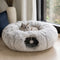 Tanière pour chat grise en peluche ultra douce avec tunnel intégré et pompon suspendu, placée près d’une fenêtre avec un chat allongé dessus