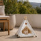 Tipi pour chat blanc avec coussin moelleux et pompons décoratifs, installé en extérieur avec un chat roux allongé dedans