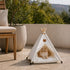Tipi pour chat blanc avec coussin moelleux et pompons décoratifs, installé en extérieur avec un chat roux allongé dedans